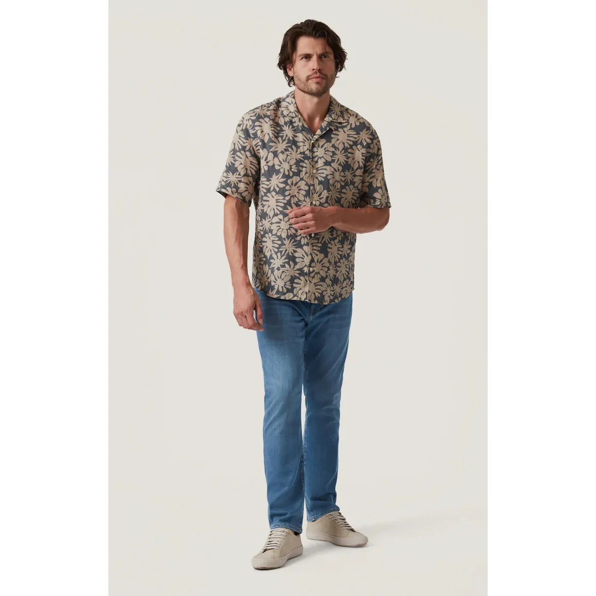 Man wearing 34 Heritage Bloom short-sleeve linen shirt with beige floral print, blue jeans, and tan sneakers.
