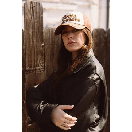 Woman in black leather jacket wearing Coors & Cowboys trucker hat with embroidered logo against rustic wooden fence.