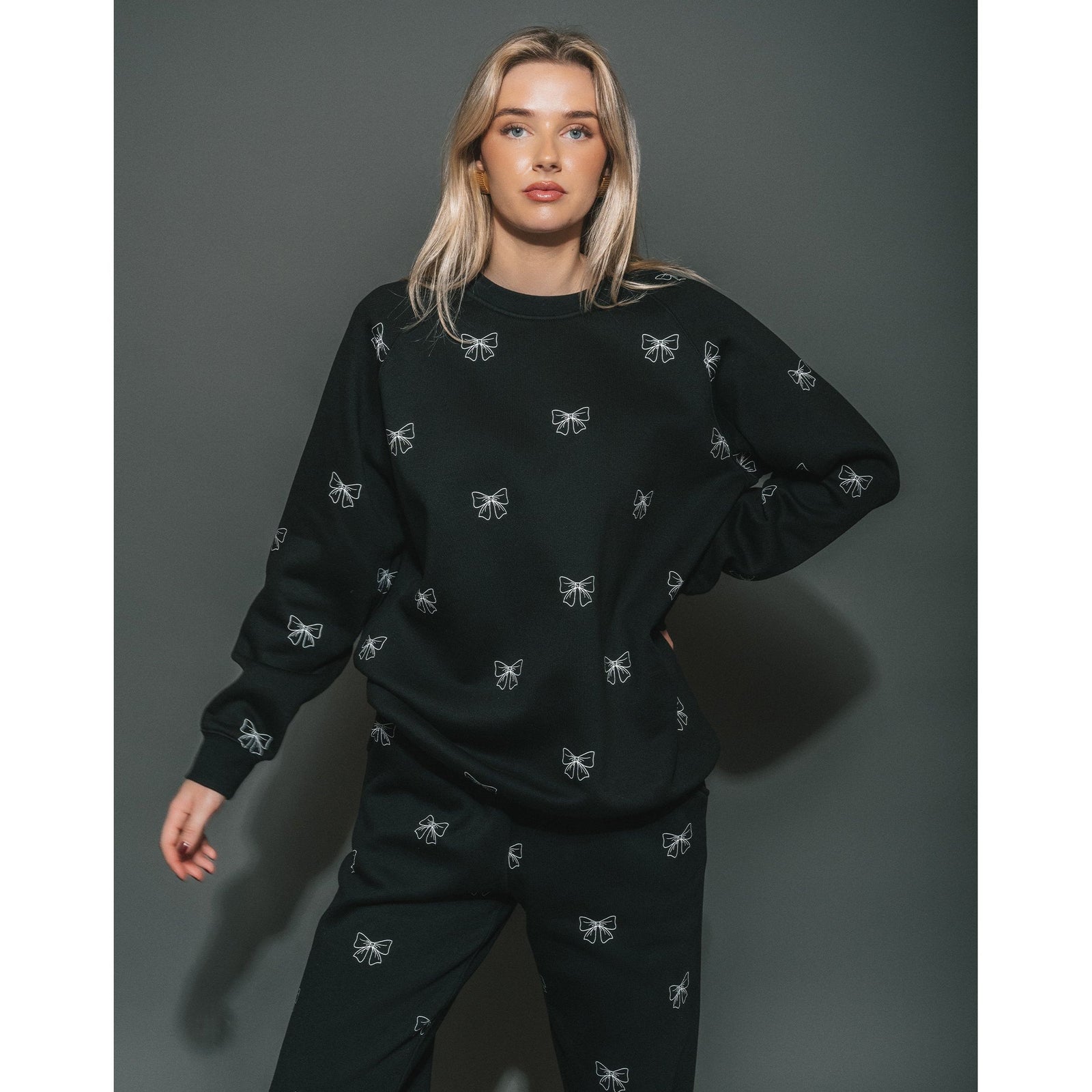 Model in Brunette the Label All Over Bow Big Sister Crew black oversized sweatshirt with white bow prints.