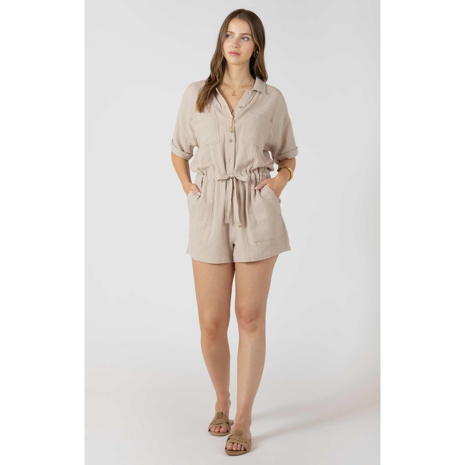 Dex Polo V-Neck Romper in beige with tied waist, button front, and side pockets on a model in sandals.