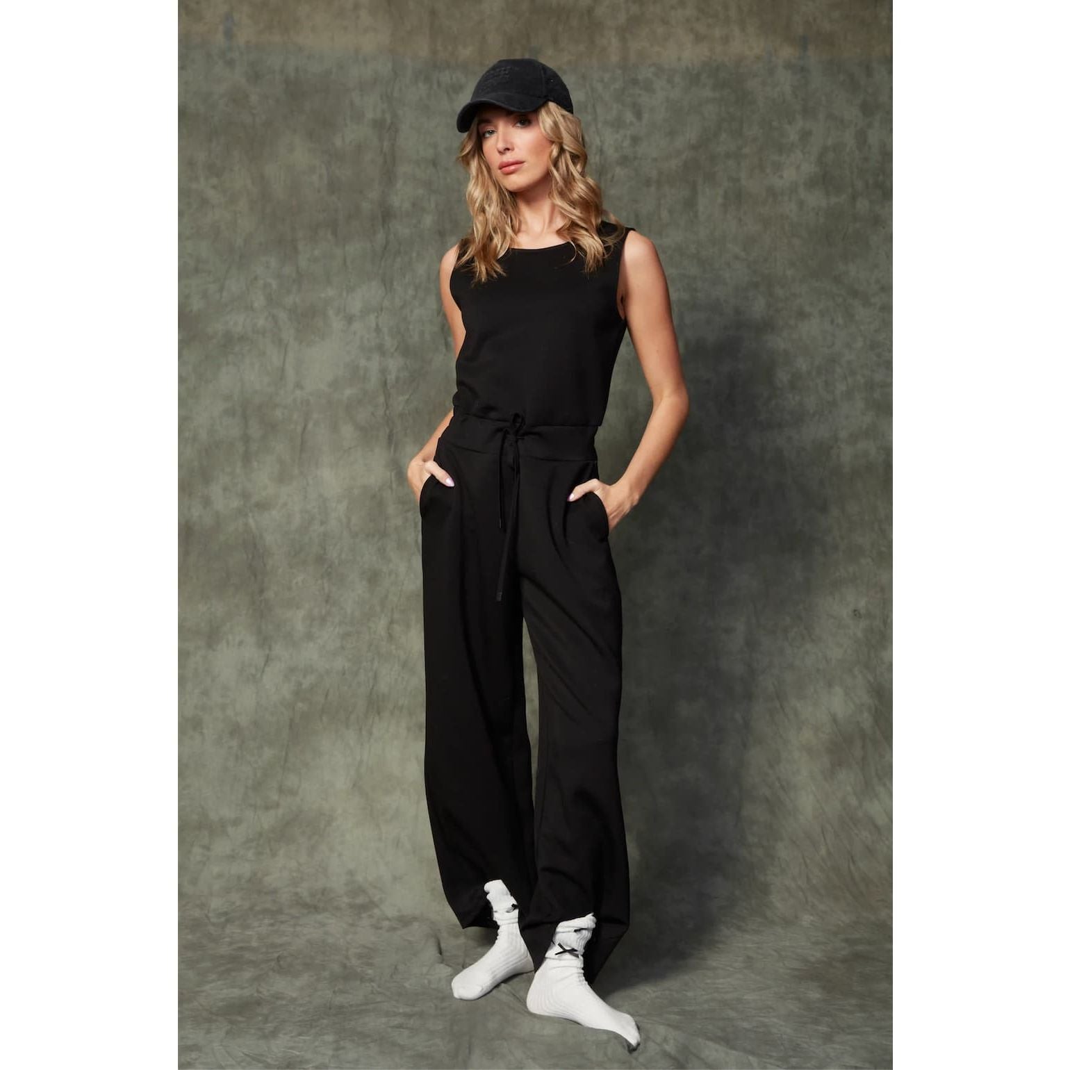 Juniva Soft Knit Jumpsuit on model: black, sleeveless, drawstring waist, wide-leg with pockets; cap and white socks.