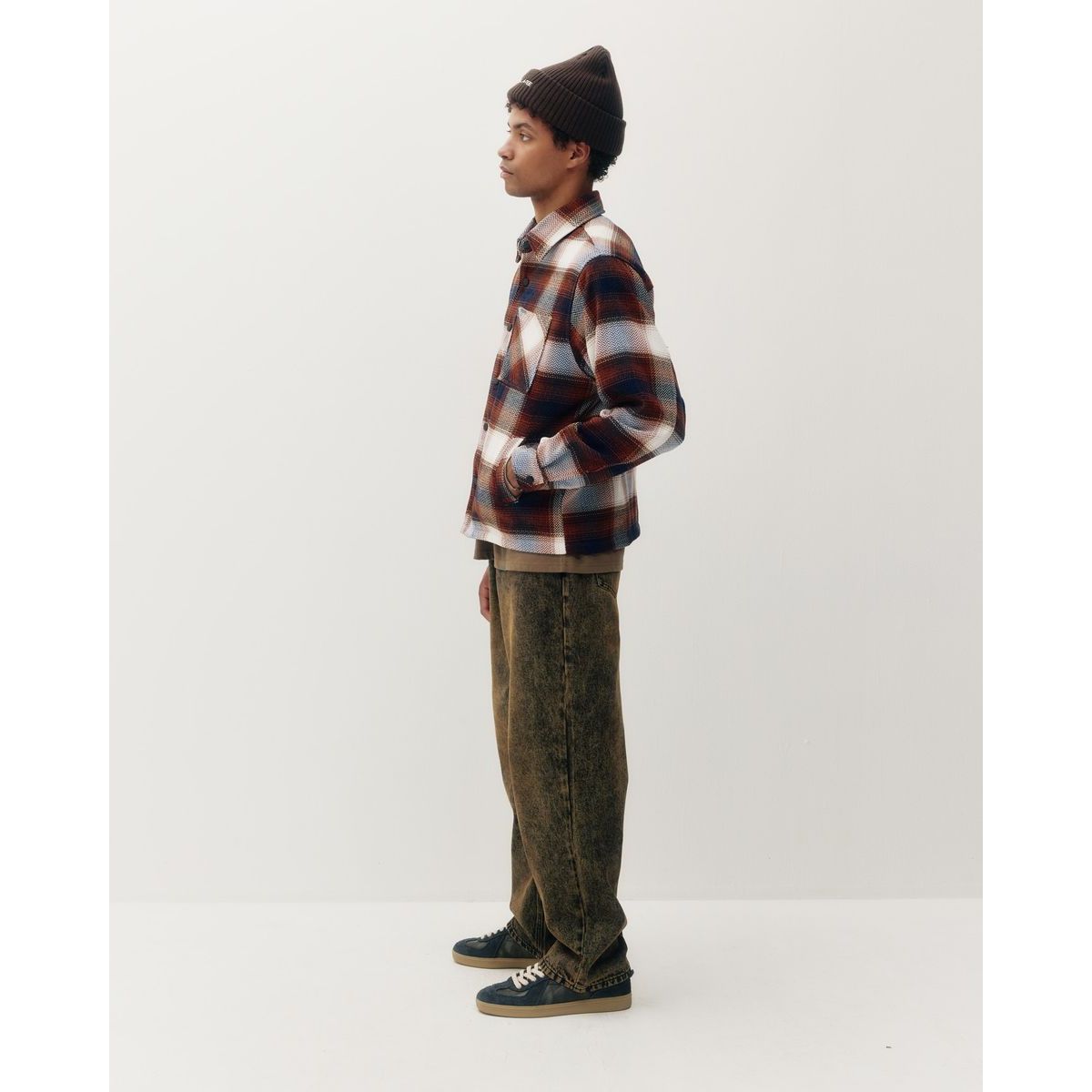 Man in side profile wearing heavyweight plaid shacket with red, blue, and white pattern, brown beanie, and faded olive jeans.