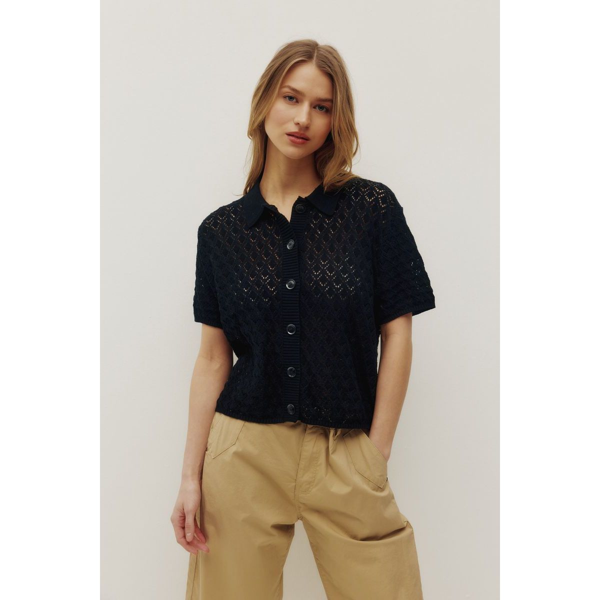 Kuwalla Monica Beach Knit Button Up in black lace-knit, short-sleeve, button-front with collar, paired with tan pants.
