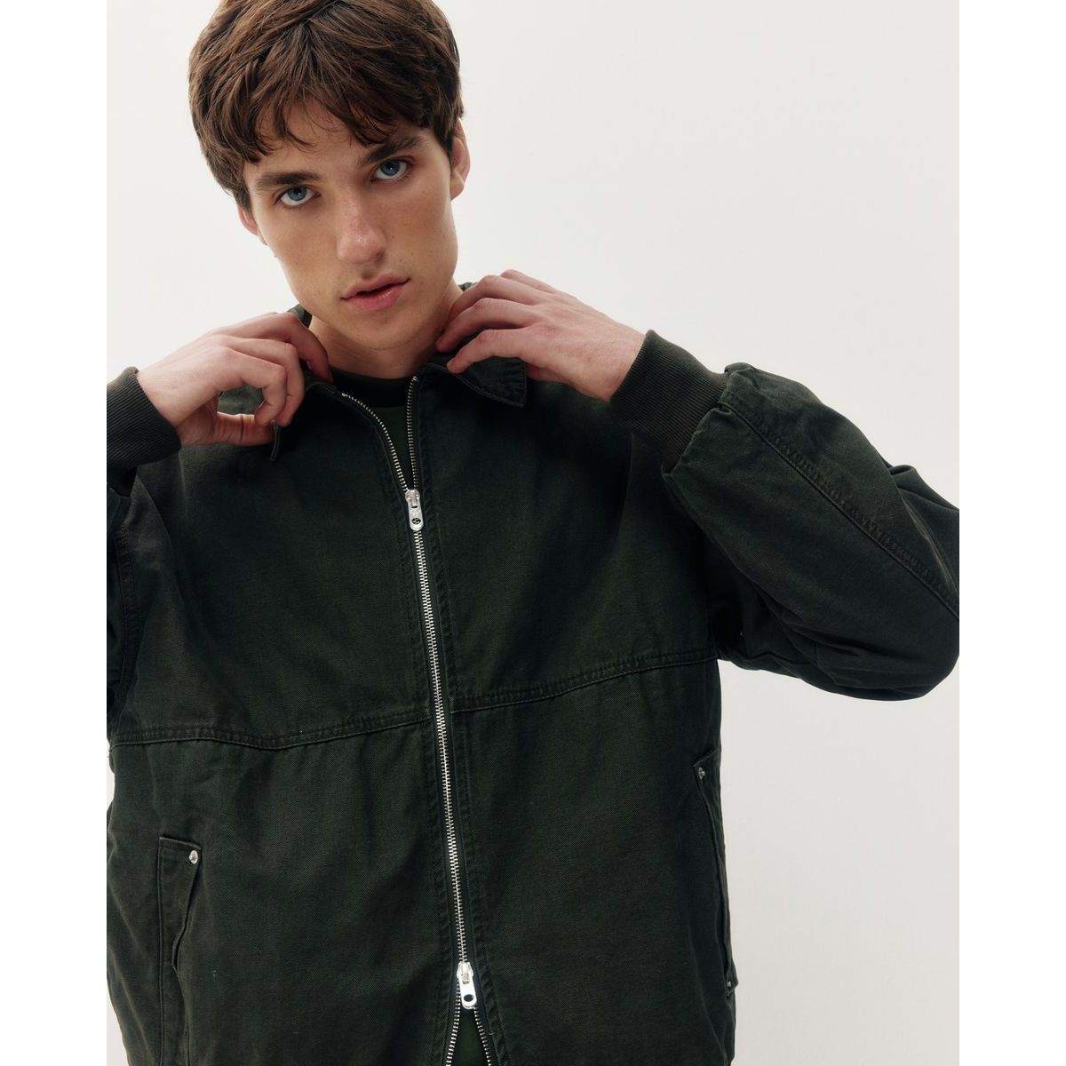 Man adjusting the structured collar of a dark green cotton Oxford bomber jacket featuring a silver zipper and minimalist utility design.