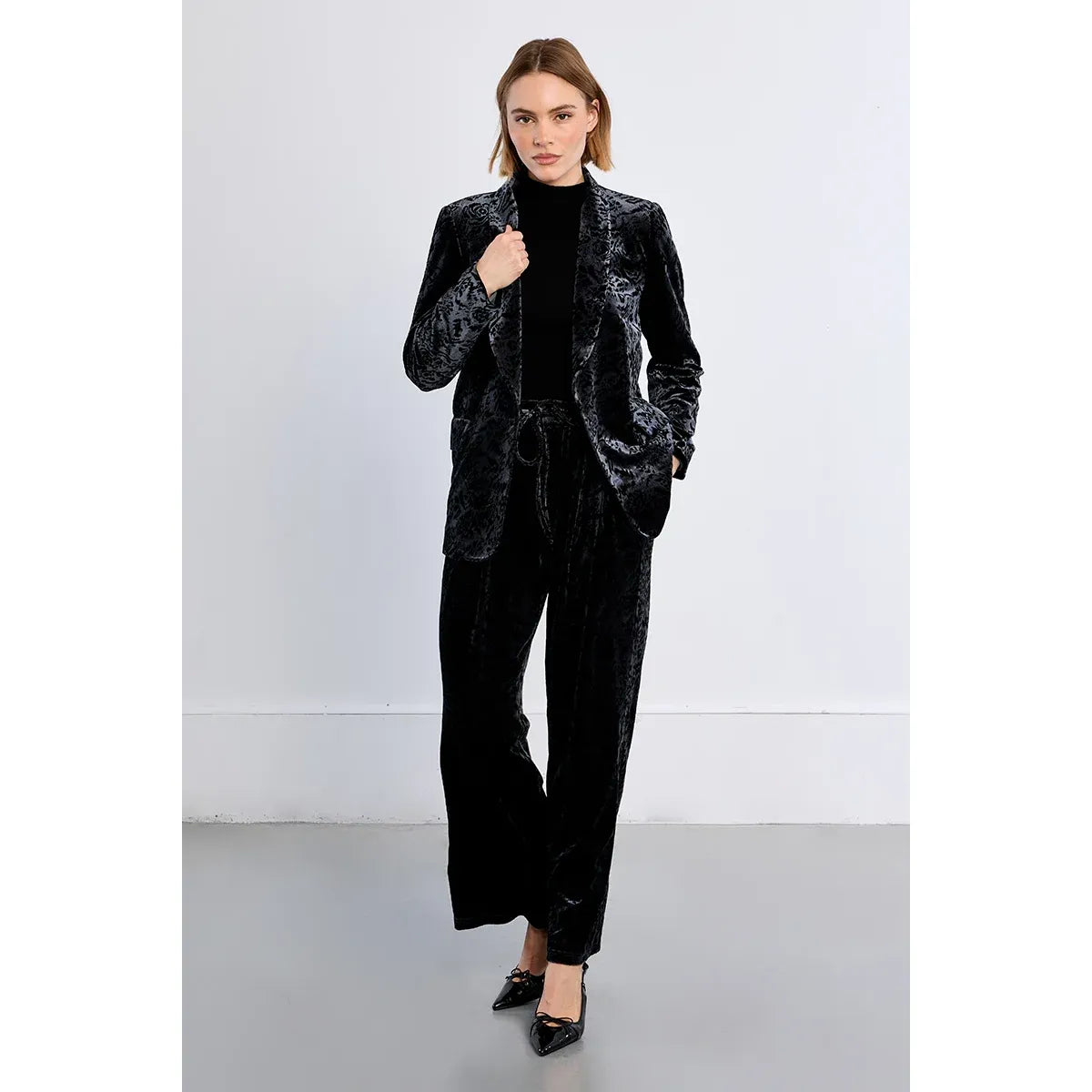Model wearing a black floral velvet blazer with a V-neck and front button closure, paired with matching velvet trousers and black heels.