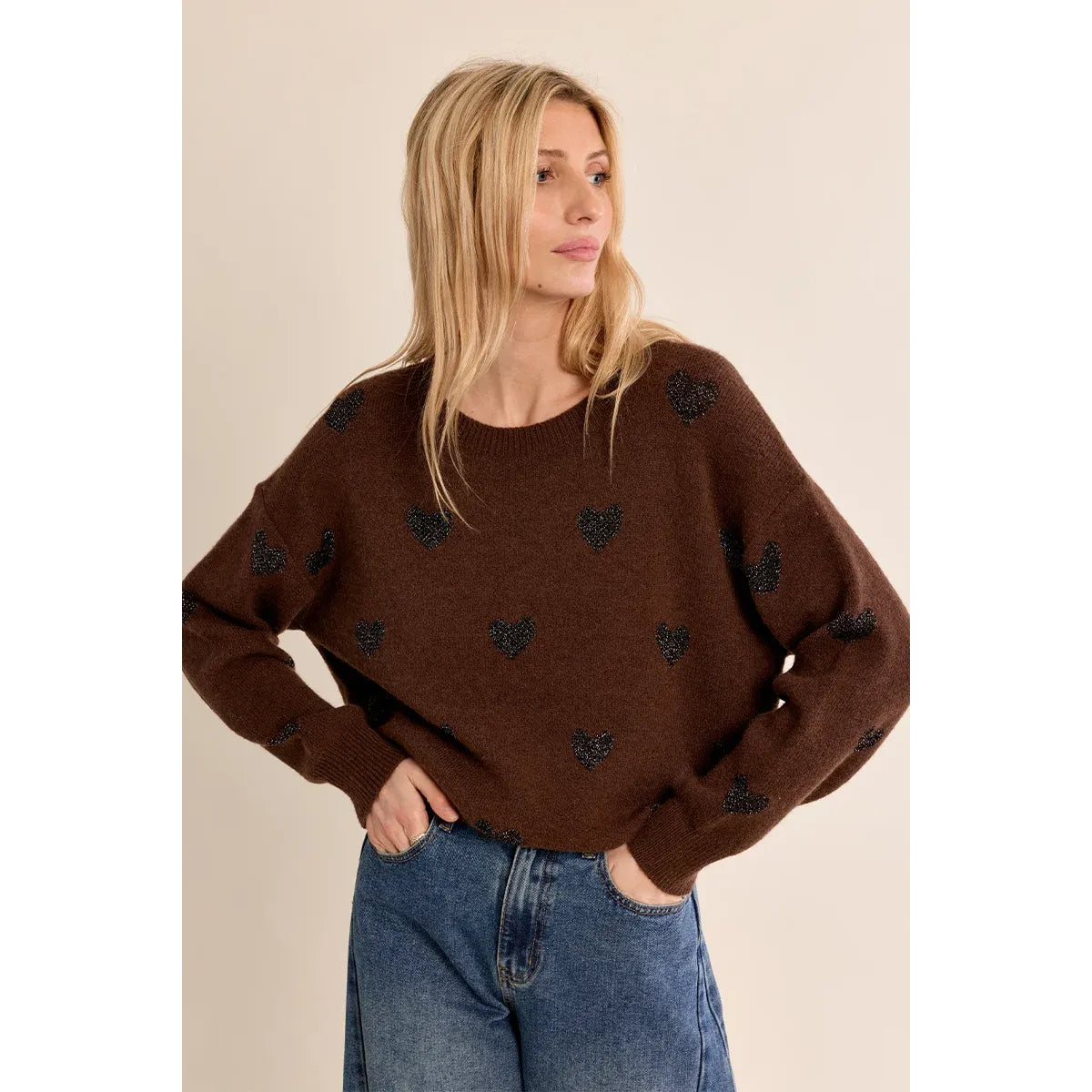 Brown knit sweater with glittery black heart pattern, round neckline, relaxed fit, and long sleeves paired with blue jeans.