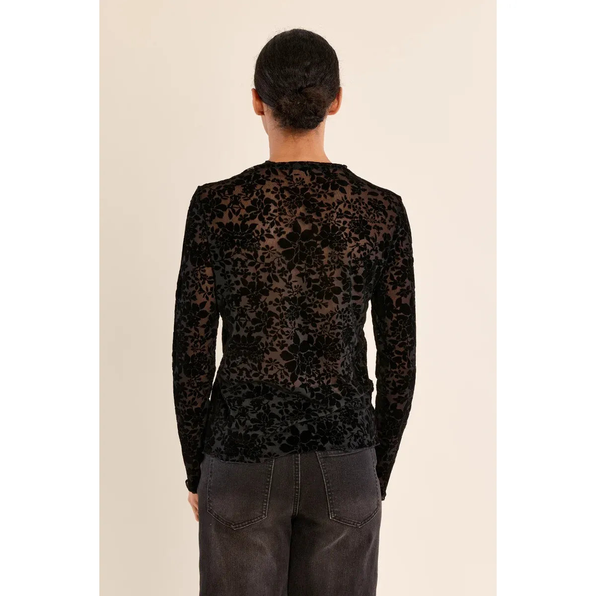 Back view of a black semi-transparent floral lace mock neck top with long sleeves, paired with dark jeans.