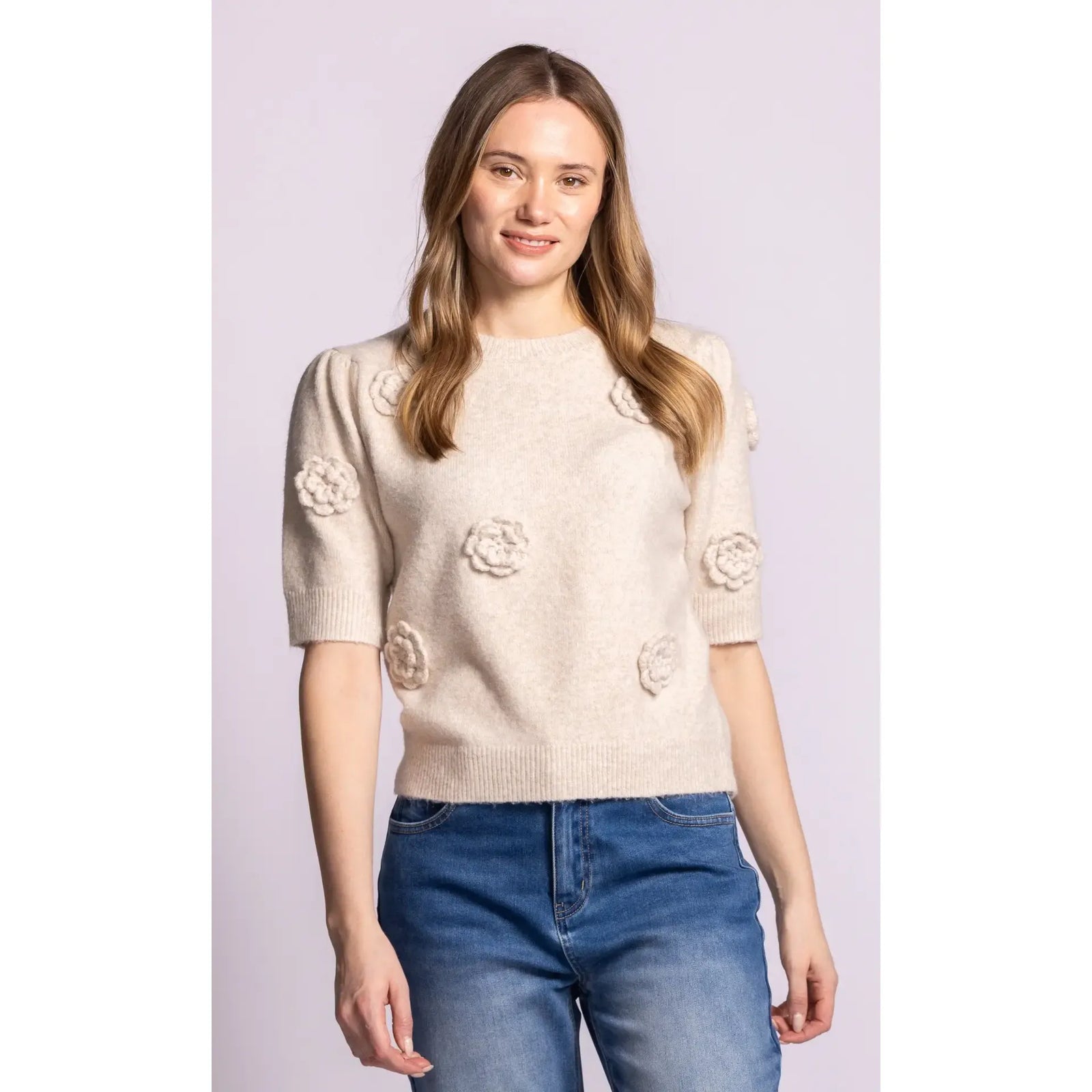 Beige knitted short-sleeve sweater with 3D rosette details and ribbed trim, paired with blue jeans for feminine charm.
