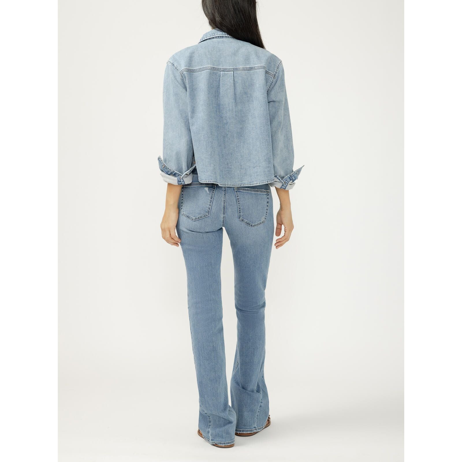 Back view of a light wash cropped denim button-up shirt with rolled sleeves and knit flex fabric, paired with flared jeans.