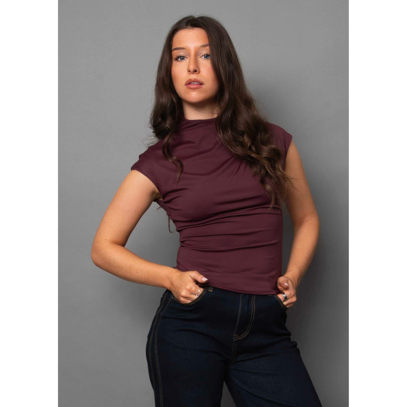Model wearing burgundy Yvette Funnel Neck Draped Top with ruched drape and cap sleeves, paired with dark jeans.