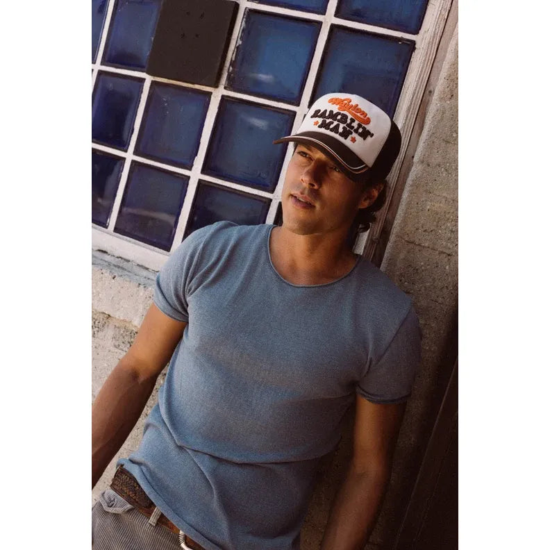 Man wearing a Waylon Jennings Radler Trucker hat with bold "Ramblin' Man" embroidery against a blue glass block wall.