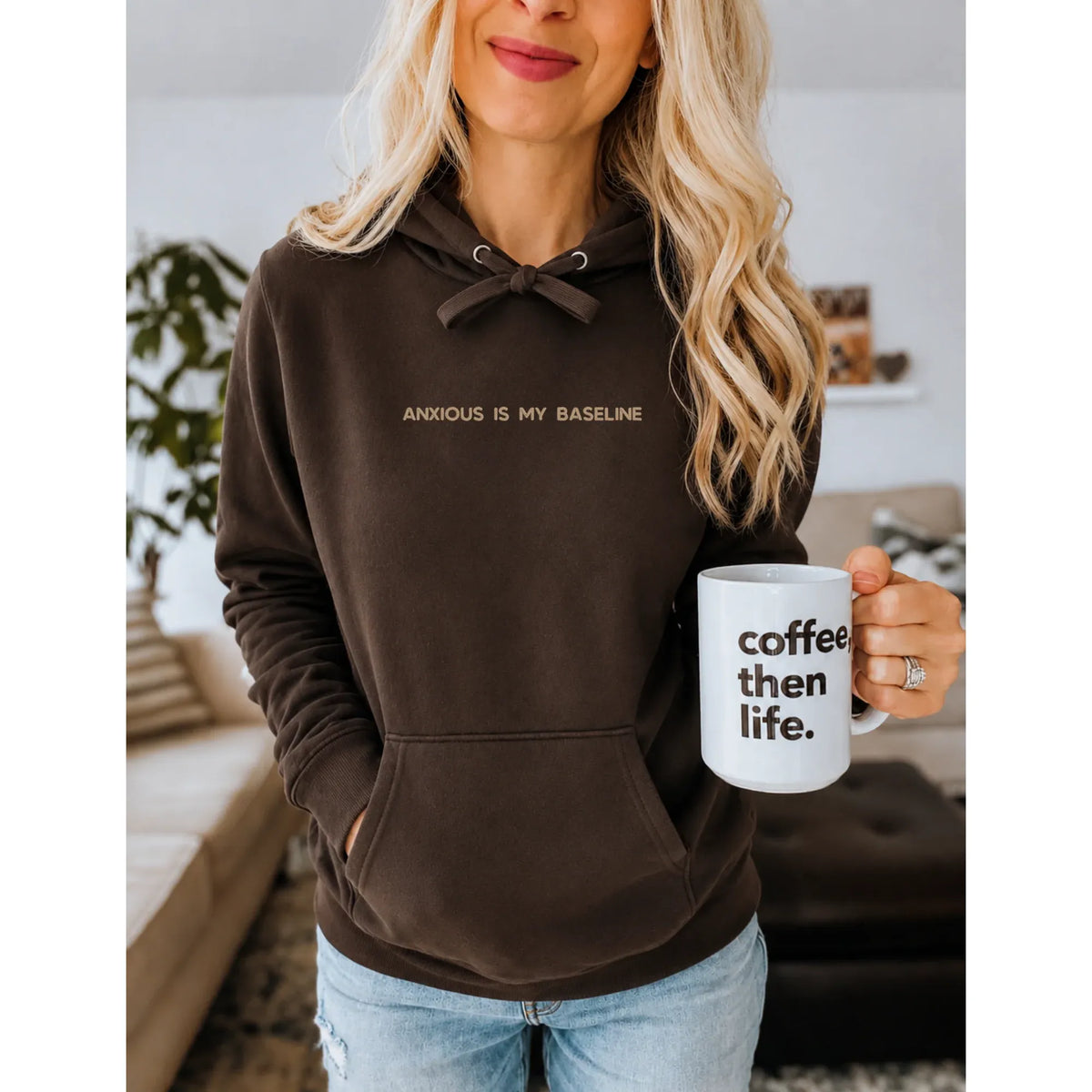 Brown embroidered hoodie reading "Anxious Is My Baseline" with bow tie drawstrings, worn while holding a coffee mug.