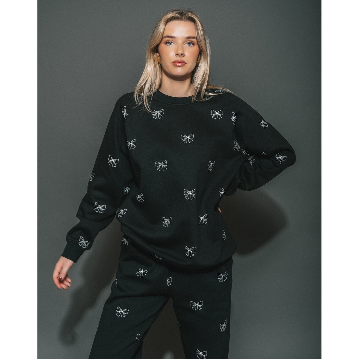 Model in Brunette the Label All Over Bow Big Sister Crew black oversized sweatshirt with white bow prints.