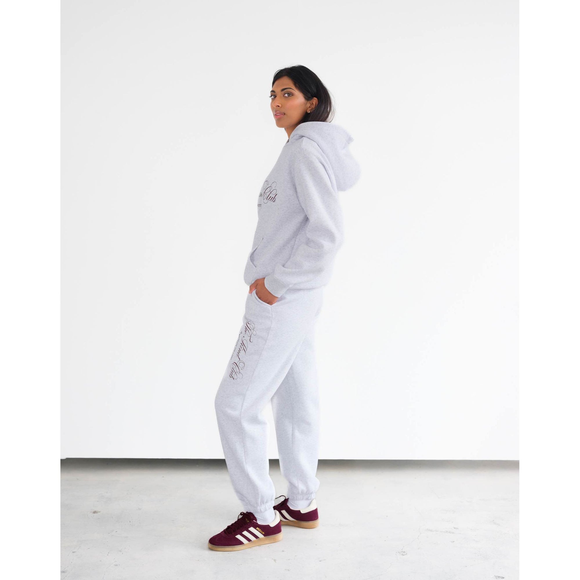 Woman wearing Brunette the Label Kind Club Core Hoodie in light gray with burgundy script on chest, paired with joggers.