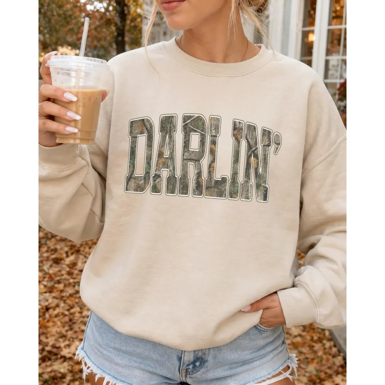 Beige Darlin' Cozy Crew sweatshirt with camo print lettering, worn with denim shorts while holding an iced coffee among autumn leaves.