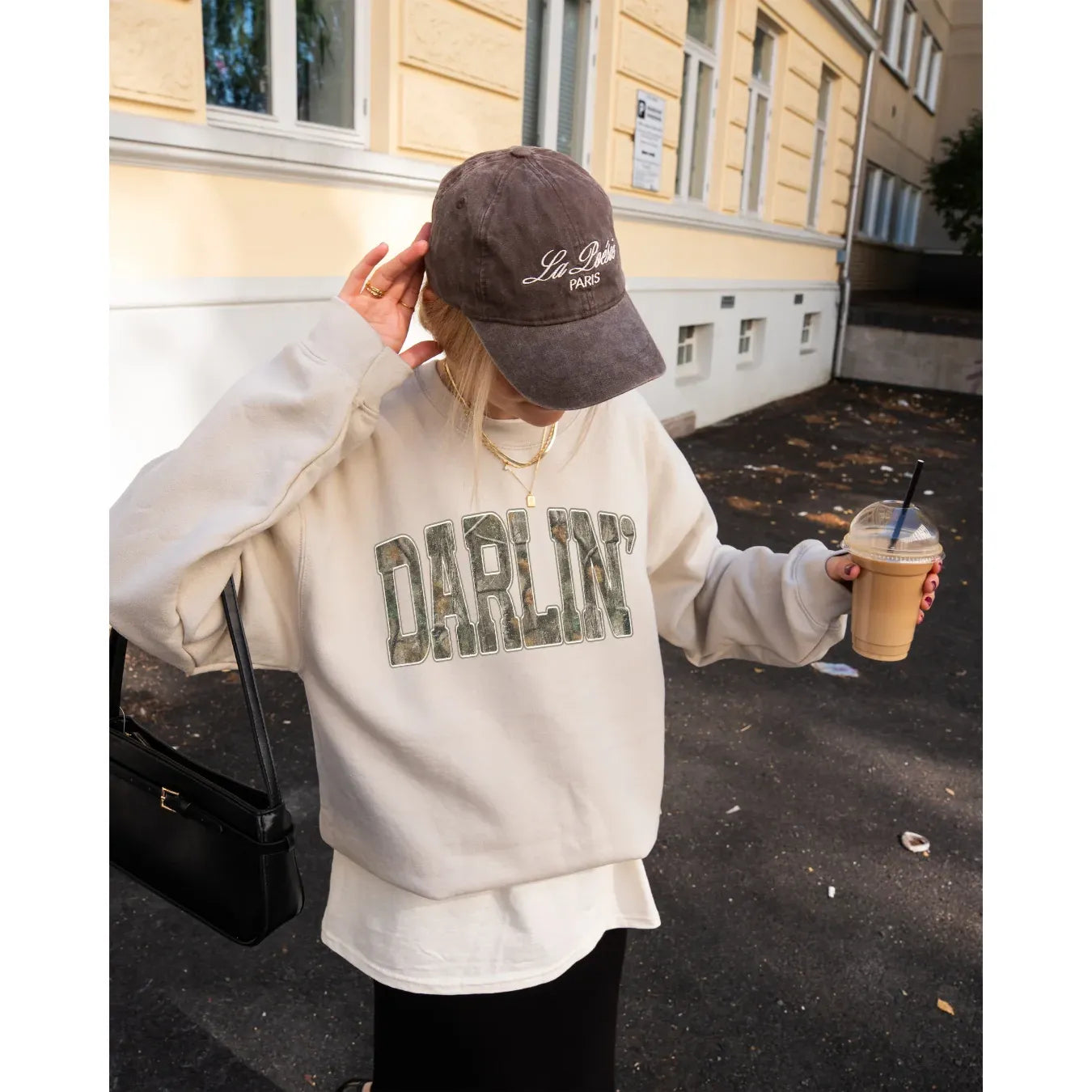 Beige Darlin' Cozy Crew sweatshirt with camo print lettering, worn with denim shorts while holding an iced coffee among autumn leaves.