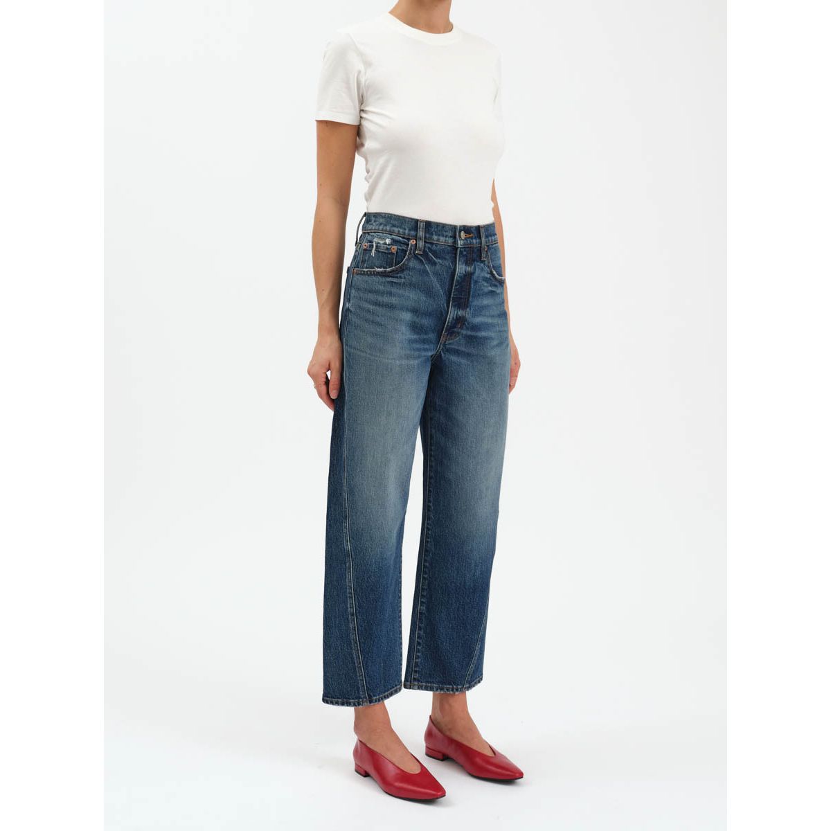 High-rise barrel-leg jeans in dark indigo with a cinched waist, structural bowed leg, and tapered ankle paired with red flats.