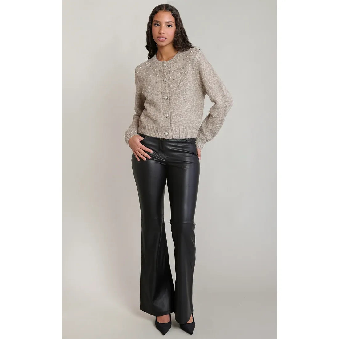 Beige embellished crewneck cardigan with pearl details and buttons, styled with black flared leather pants and heels.