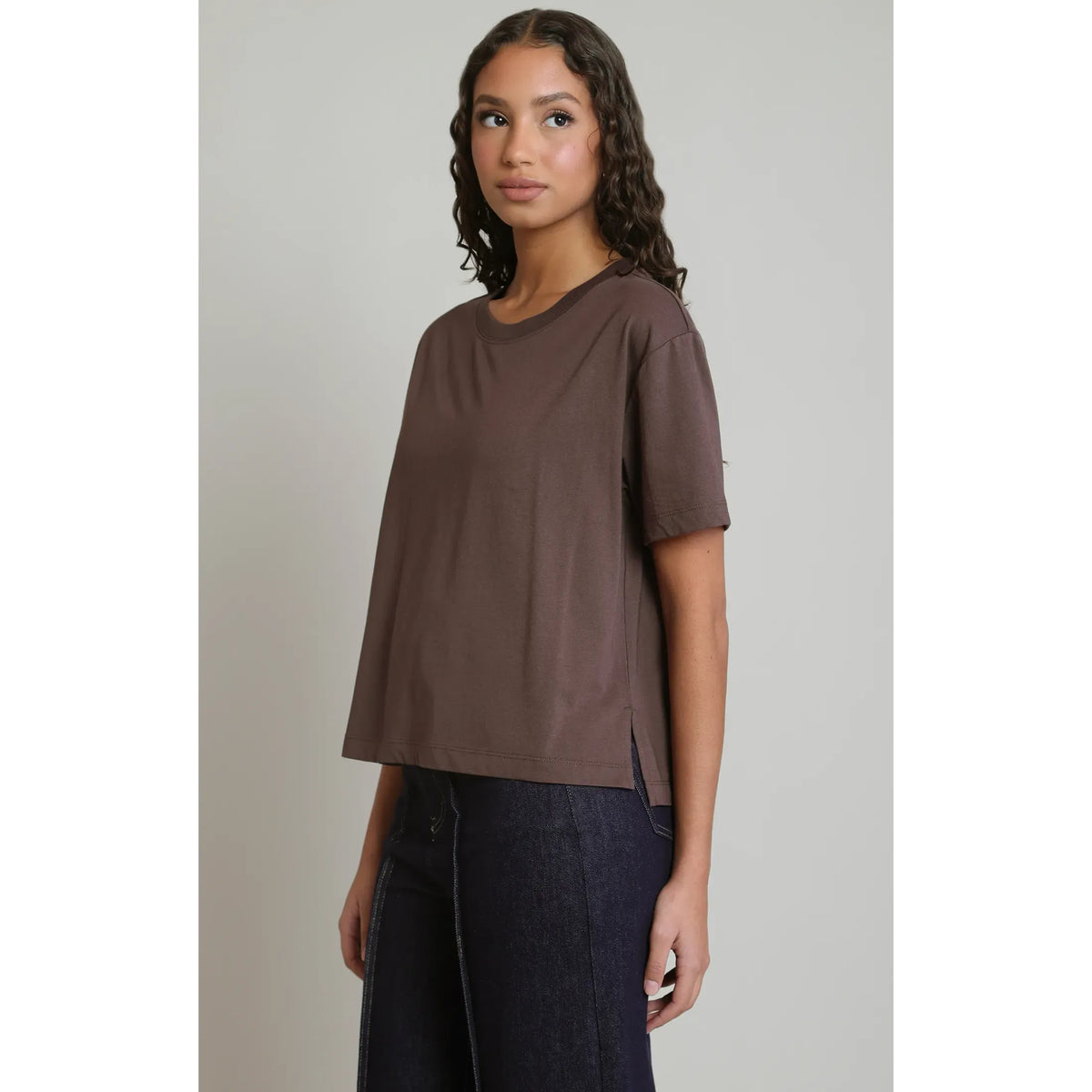 Model wearing a brown short-sleeved tee with a relaxed fit, round neck, and side slits paired with dark denim jeans.