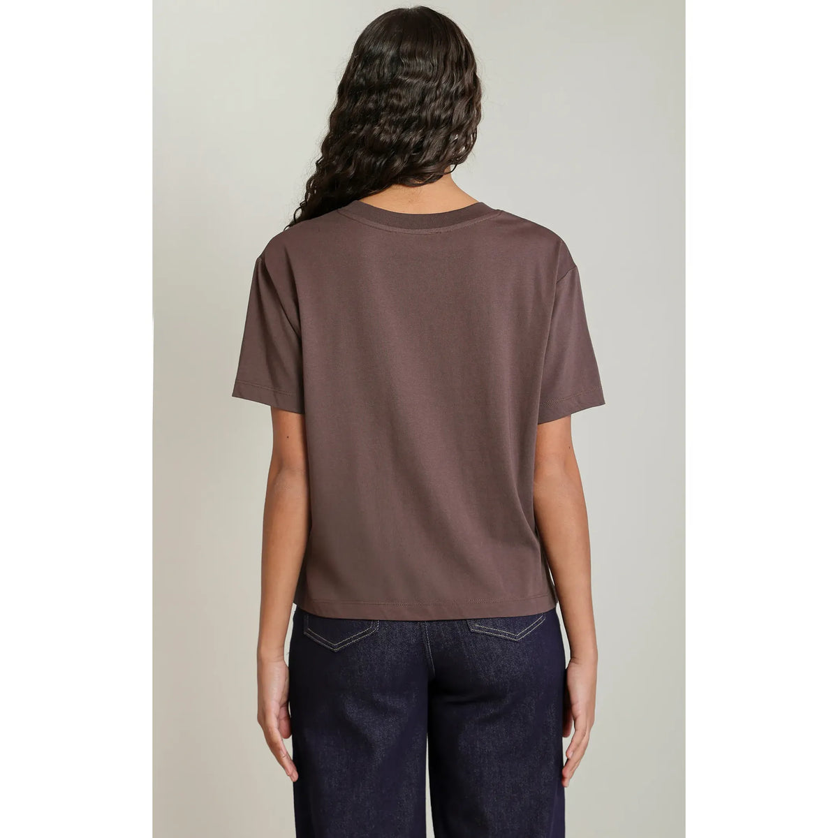 Back view of a model wearing a brown short-sleeved essential tee with a relaxed fit and round neck, paired with dark denim jeans.