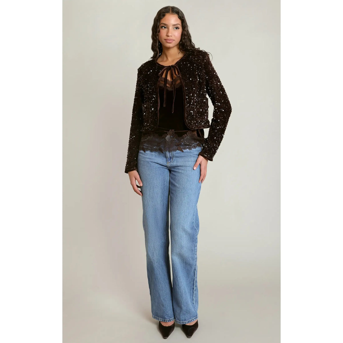 Model wearing a chocolate velvet sequin cropped jacket with lace tie neck, paired with wide-leg jeans and black heels.