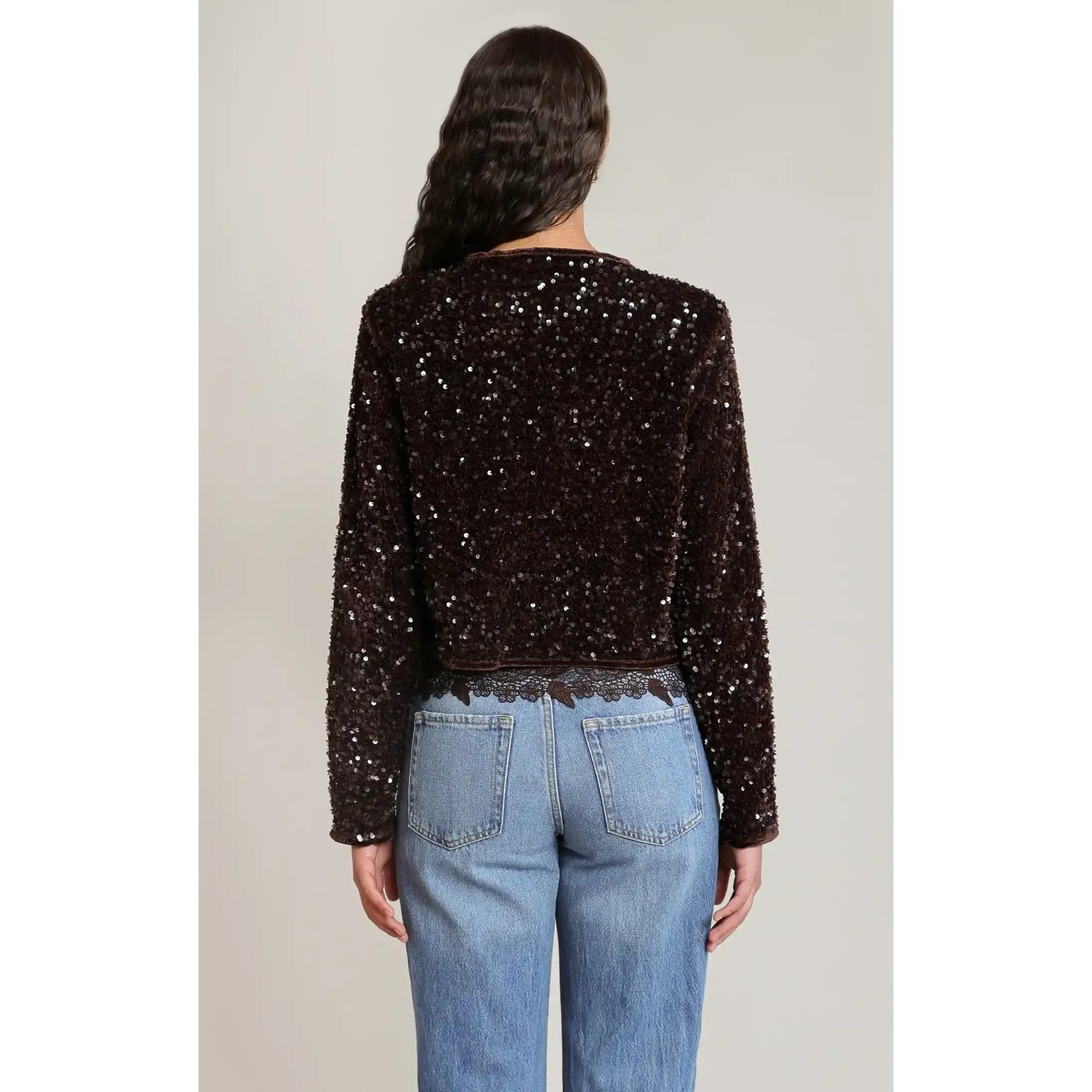 Back view of a chocolate brown cropped velvet sequin jacket with light-catching shimmer, styled with blue jeans and lace trim.