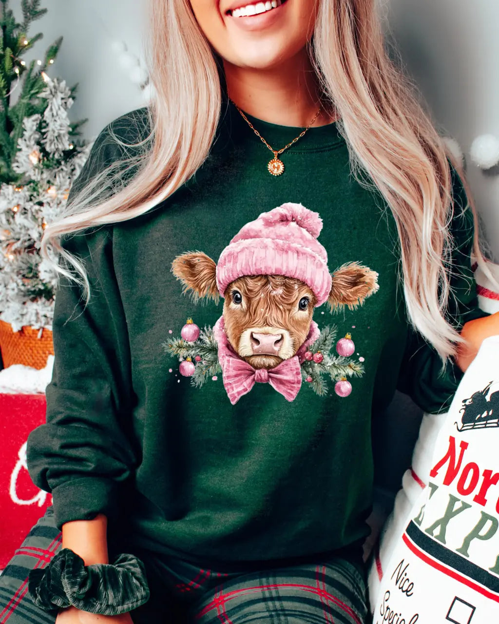 Green crewneck sweatshirt featuring a Highland calf in a pink pom-pom hat and bow tie, framed by pine sprigs and holiday ornaments.