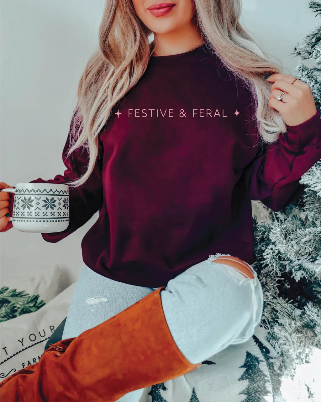 Burgundy crewneck sweatshirt printed with "Festive &amp; Feral" in pink, held with a holiday mug near a flocked tree.