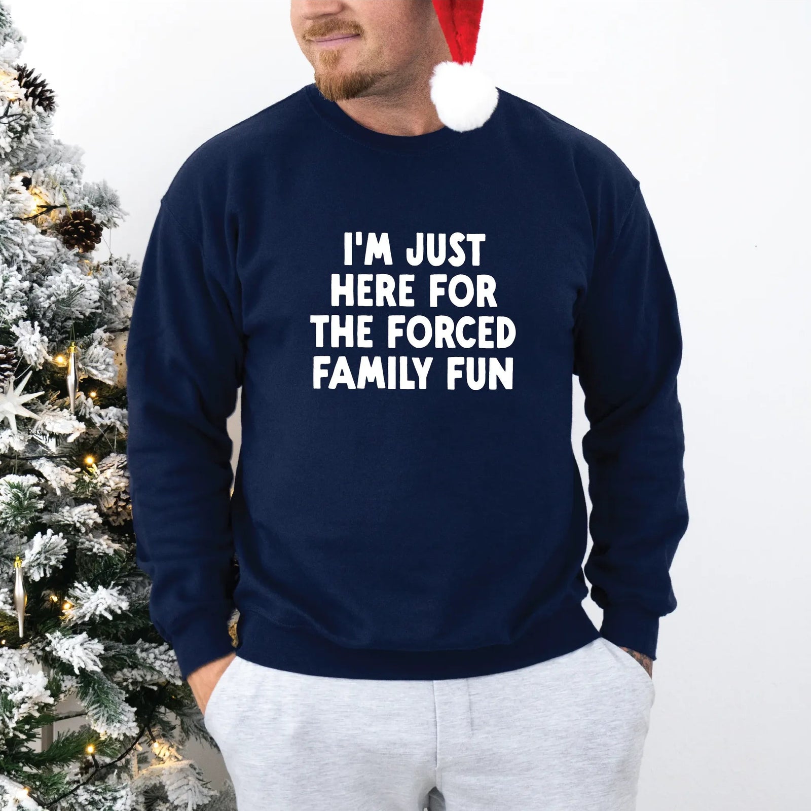 Man wearing navy Forced Family Fun Crew sweatshirt with bold white text and Santa hat beside a snowy Christmas tree.