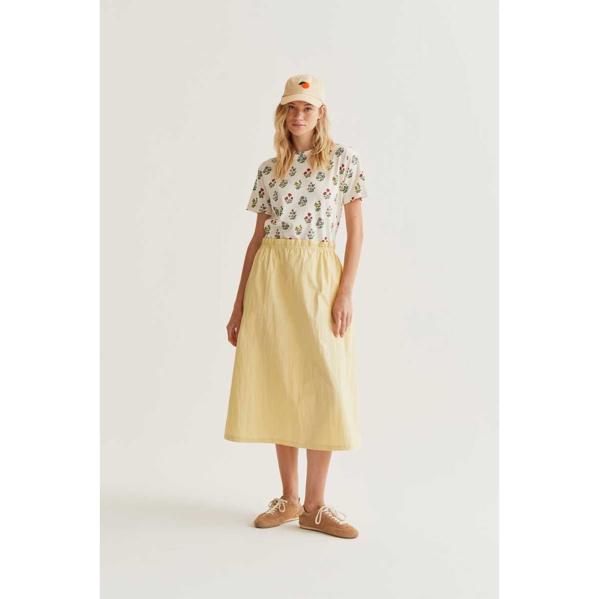 Gardener Print Tee on a model with a whimsical multi-coloured botanical print, paired with a pale yellow midi skirt and beige cap.
