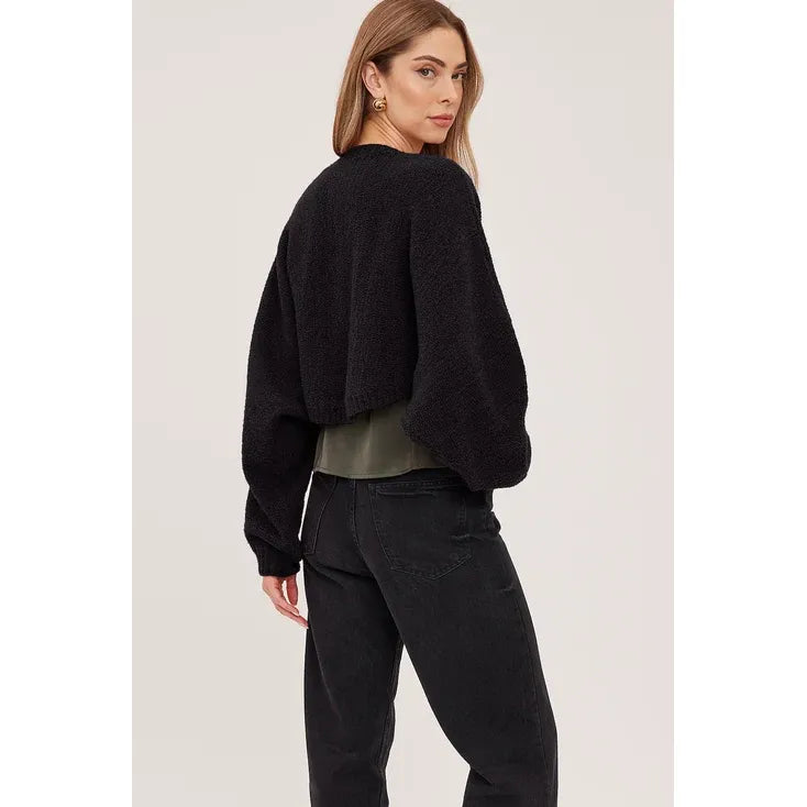 Black cropped open-front bouclé cardigan with voluminous sleeves and fitted rib cuffs, styled over an olive top and black jeans.