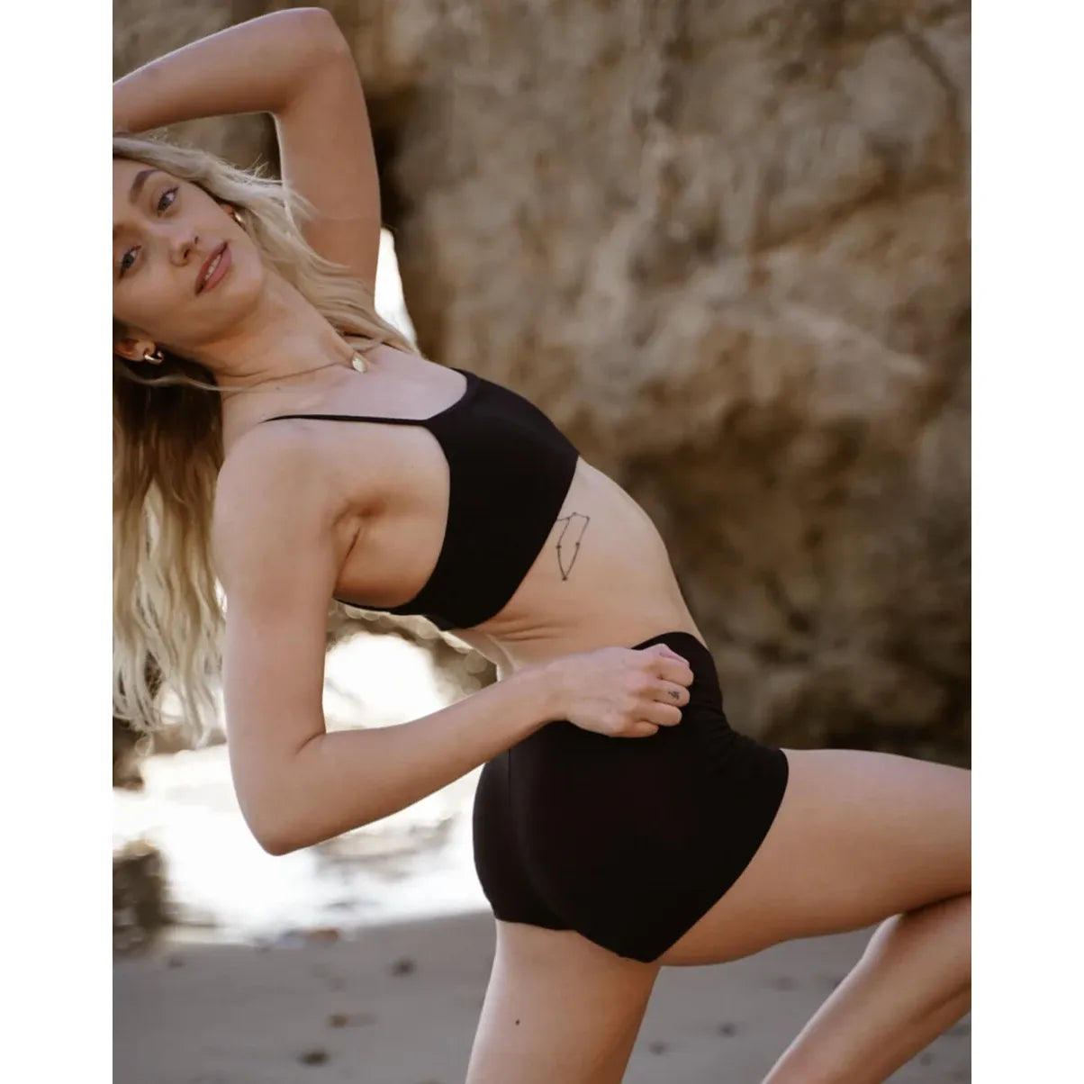 Woman wearing a black Huha Tata Mineral Bralette on the beach; hardware-free bandeau with elastic straps.