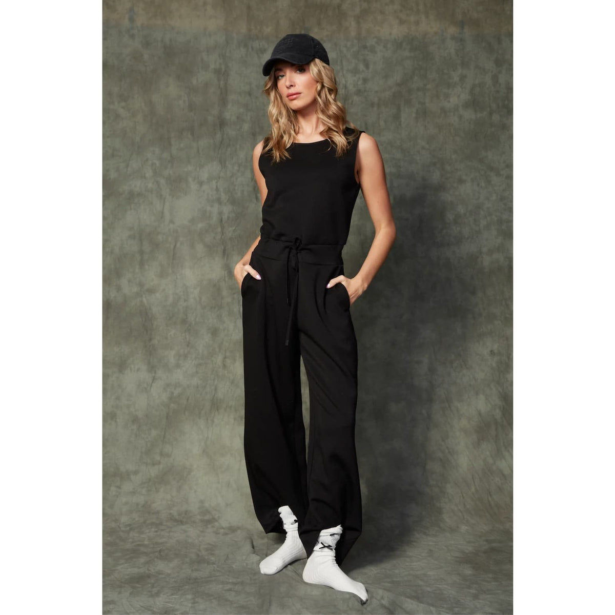 Juniva Soft Knit Jumpsuit on model: black, sleeveless, drawstring waist, wide-leg with pockets; cap and white socks.