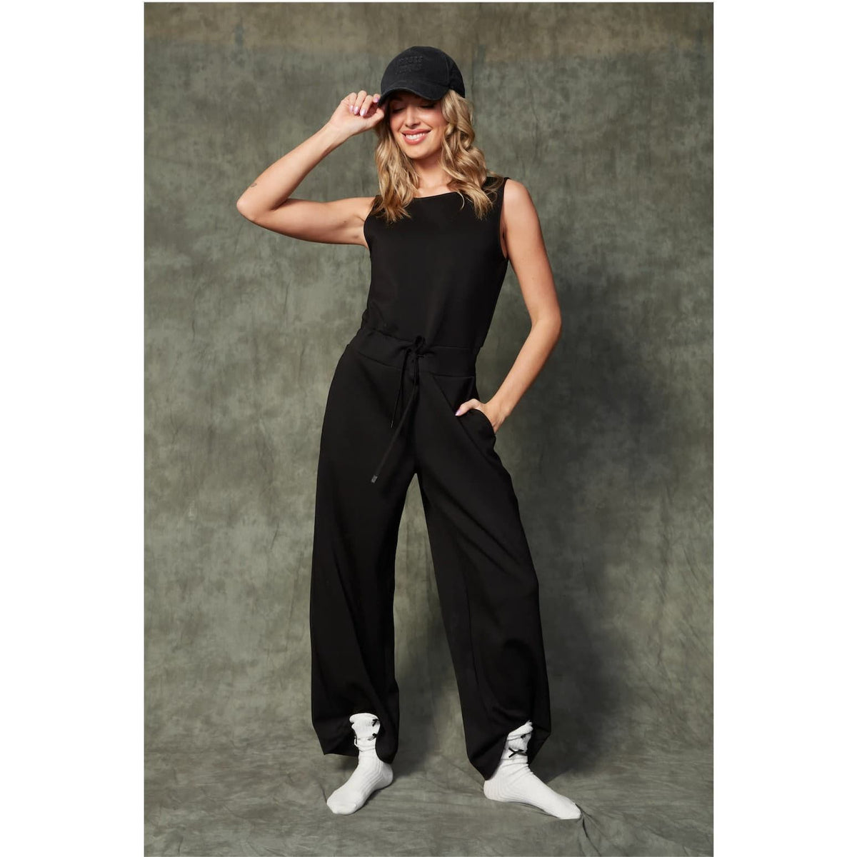 Model wearing Juniva Soft Knit Jumpsuit: sleeveless black jumpsuit with waist tie and wide-leg, ankle-length.