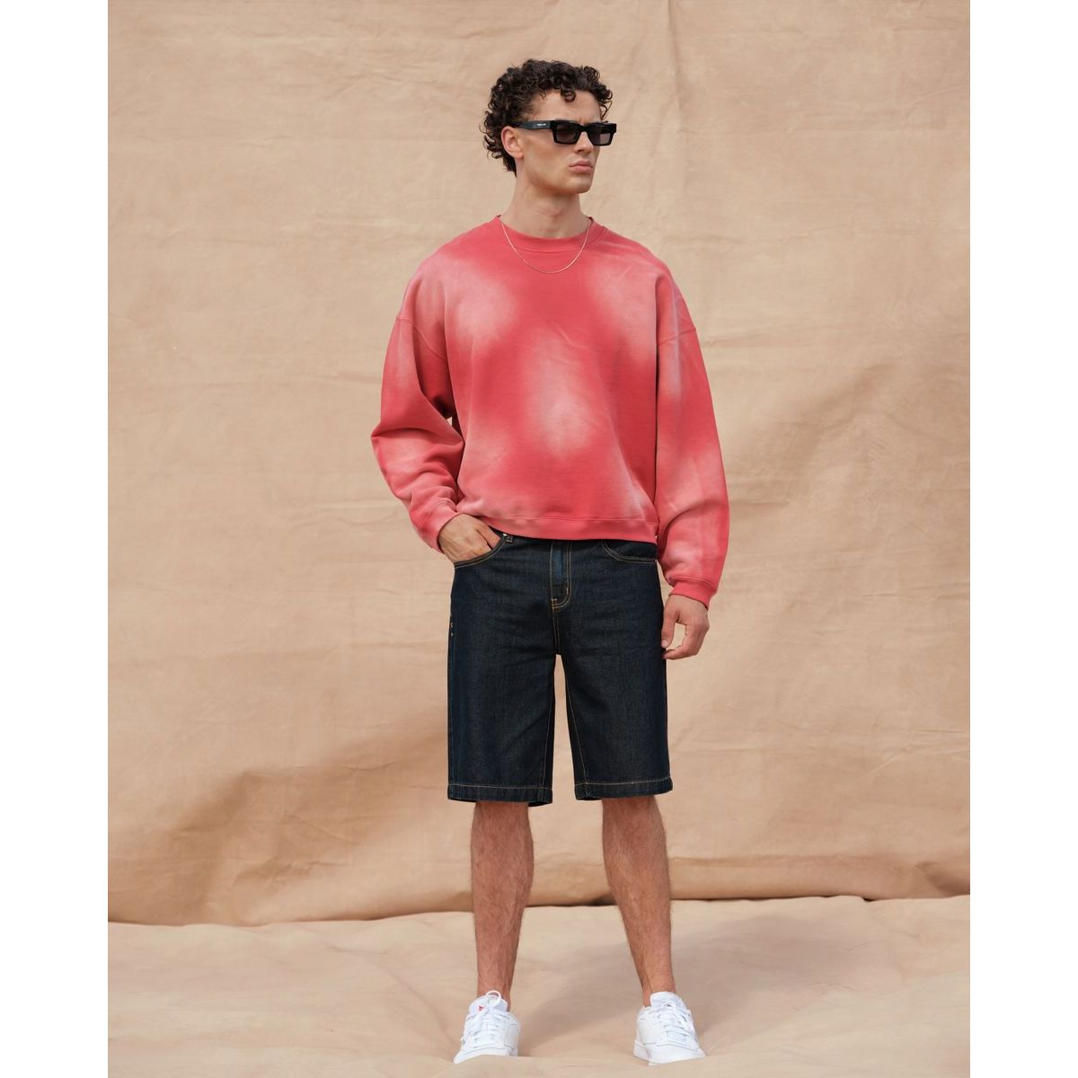 Kuwalla Bermuda Denim Shorts in dark denim, knee-length, worn by a man in a pink sweatshirt and white sneakers.