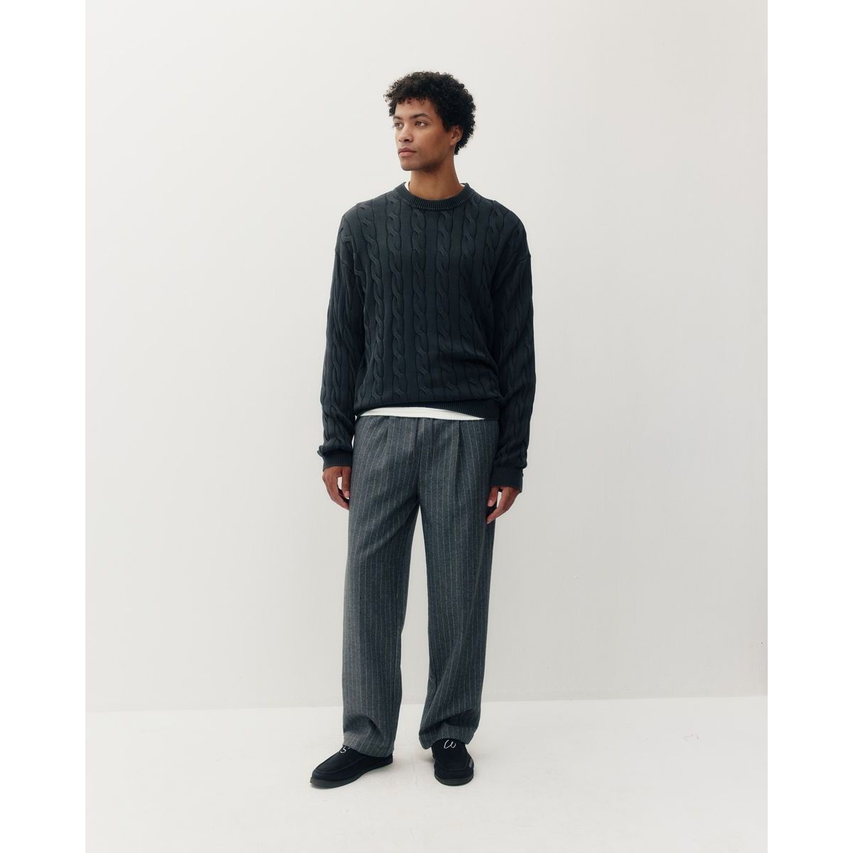 Man wearing a black braided knit box crew sweater and grey pinstripe pants, shown in profile against a white background.