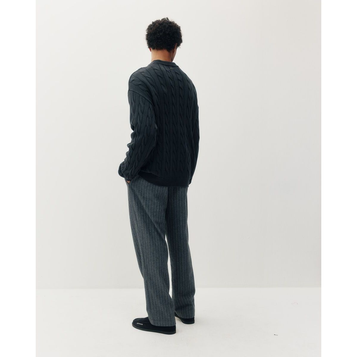 Man wearing a black braided knit box crew sweater and grey pinstripe pants, shown in profile against a white background.