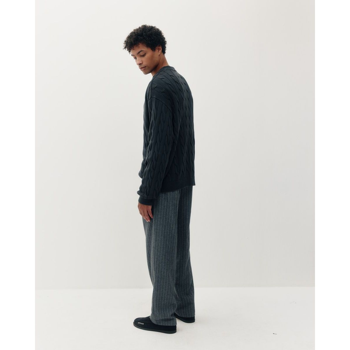 Man wearing a black braided knit box crew sweater and grey pinstripe pants, shown in profile against a white background.