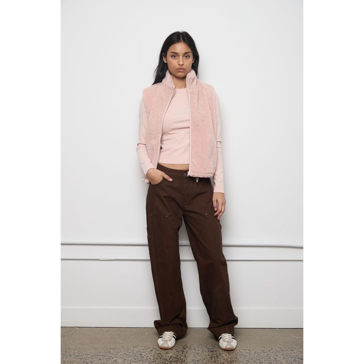 Model wearing a light pink faux fur zip-up vest over a matching top with brown trousers and sneakers against a white wall.