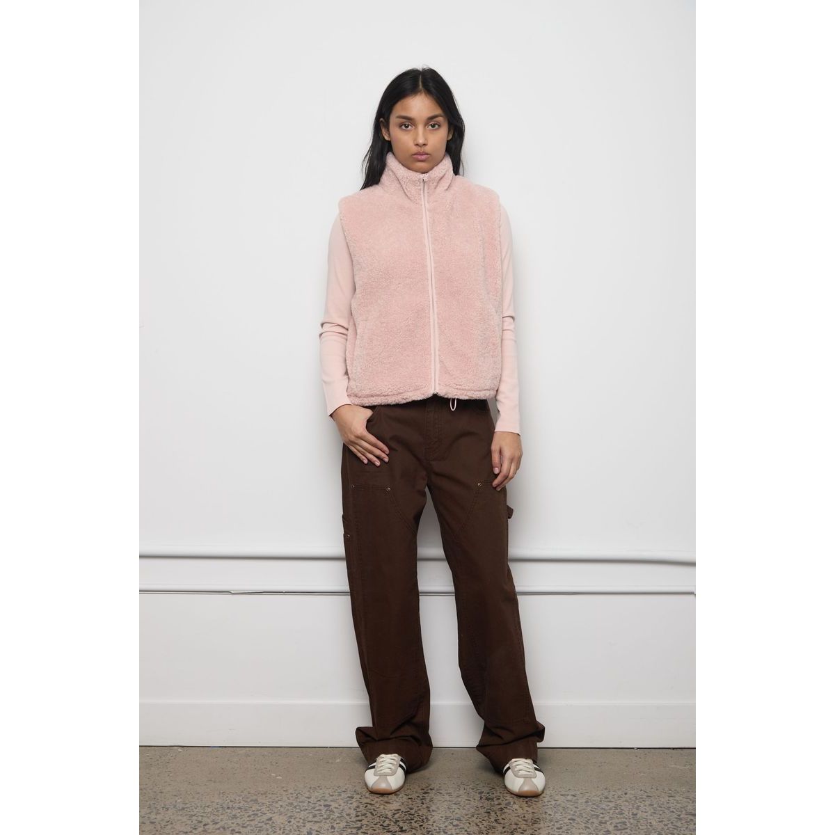 Model wearing a soft pink faux fur zip-up vest with a high collar, layered over a matching long-sleeve top and brown trousers.