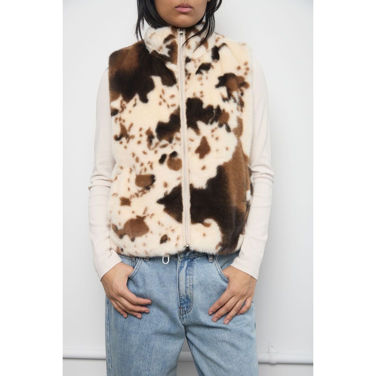 Brown and cream cow print faux fur zip-up vest worn over a fitted beige top with light blue denim jeans.