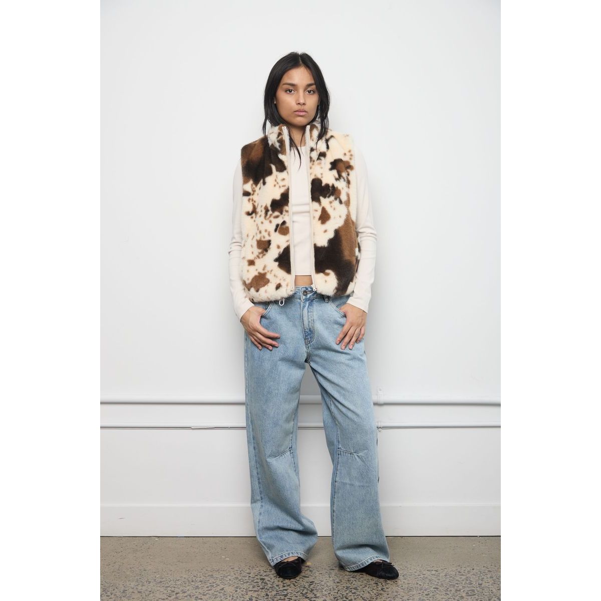 Model wears a brown and cream cow-print faux fur zip-up vest over a white top with wide-leg jeans.