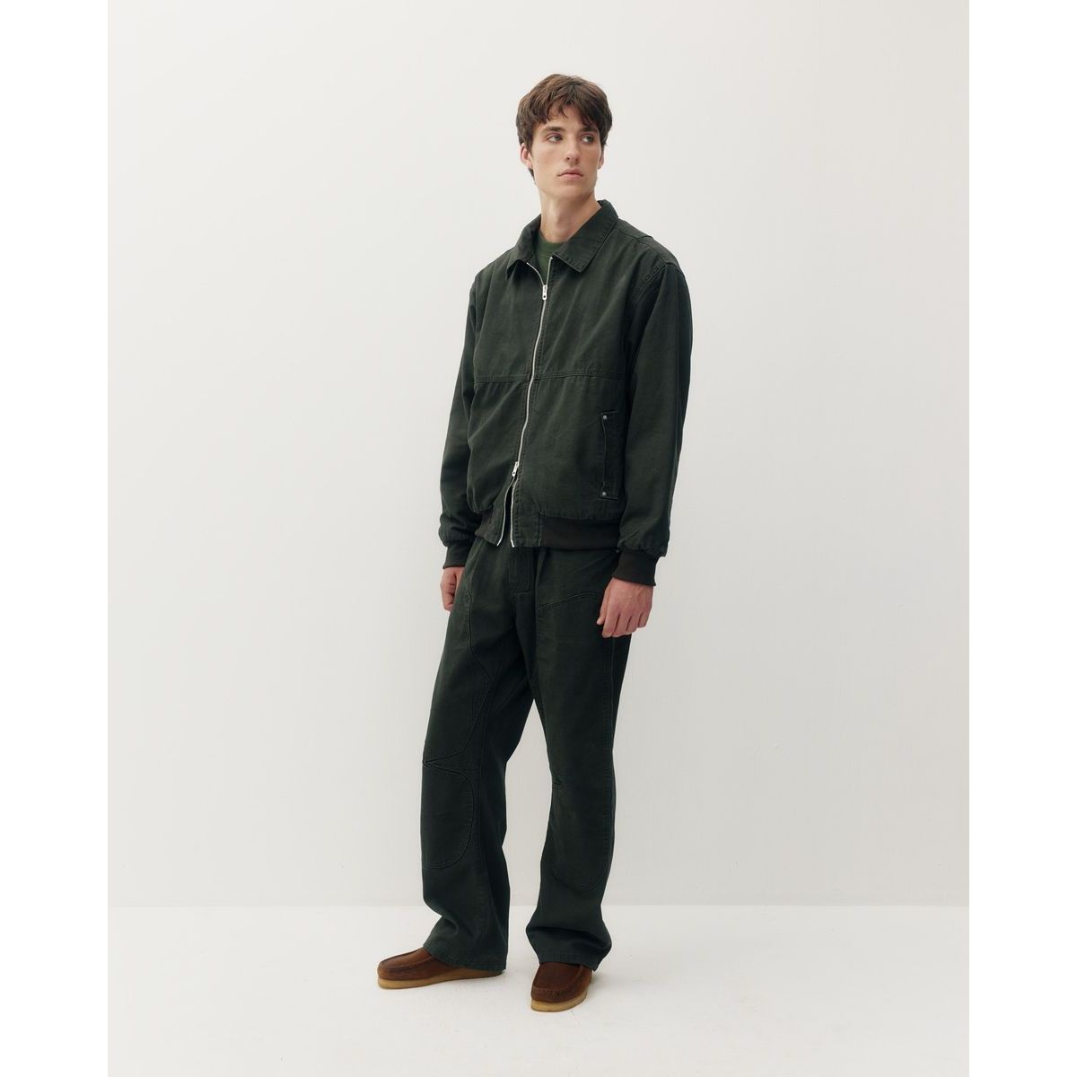 Model wears dark green Oxford Bomber jacket with structured collar and silver zipper, paired with matching cotton trousers and brown loafers.