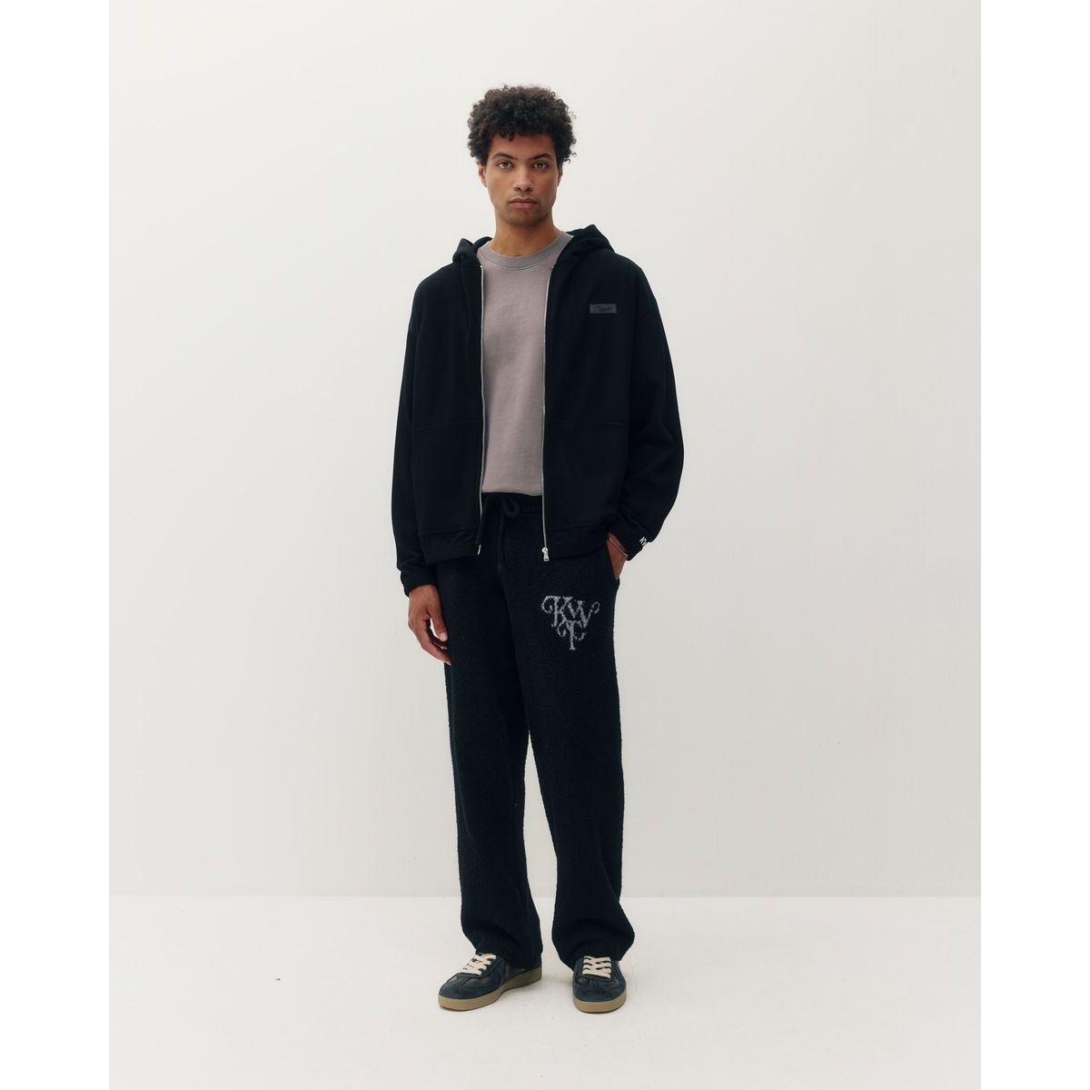 Man wearing black Kuwalla ribbed zip hoodie over grey tee with matching black sweatpants featuring white embroidered logo.