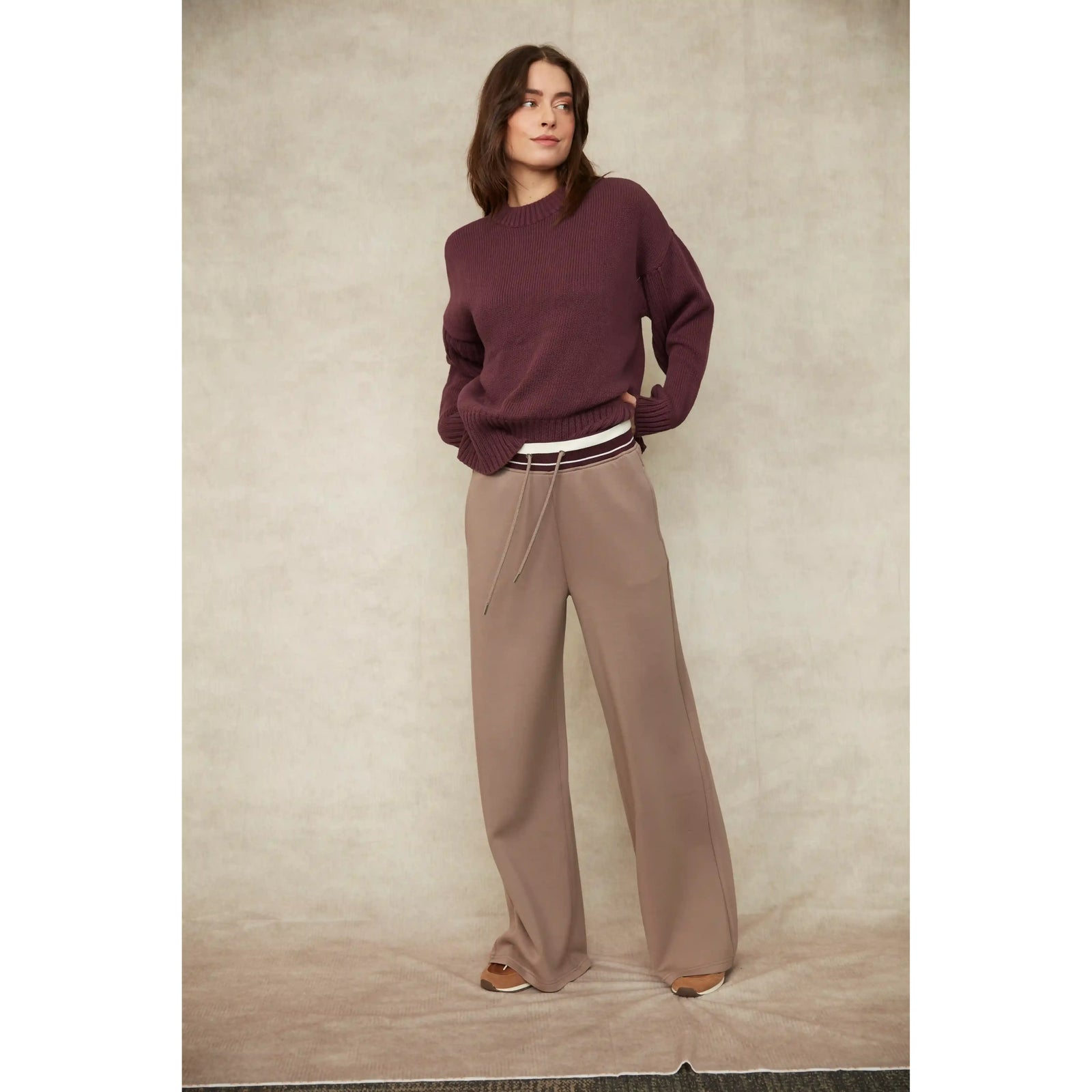 Model wearing a maroon long sleeve crew neck knit sweater with ribbed trim and side slits, paired with taupe wide-leg pants.