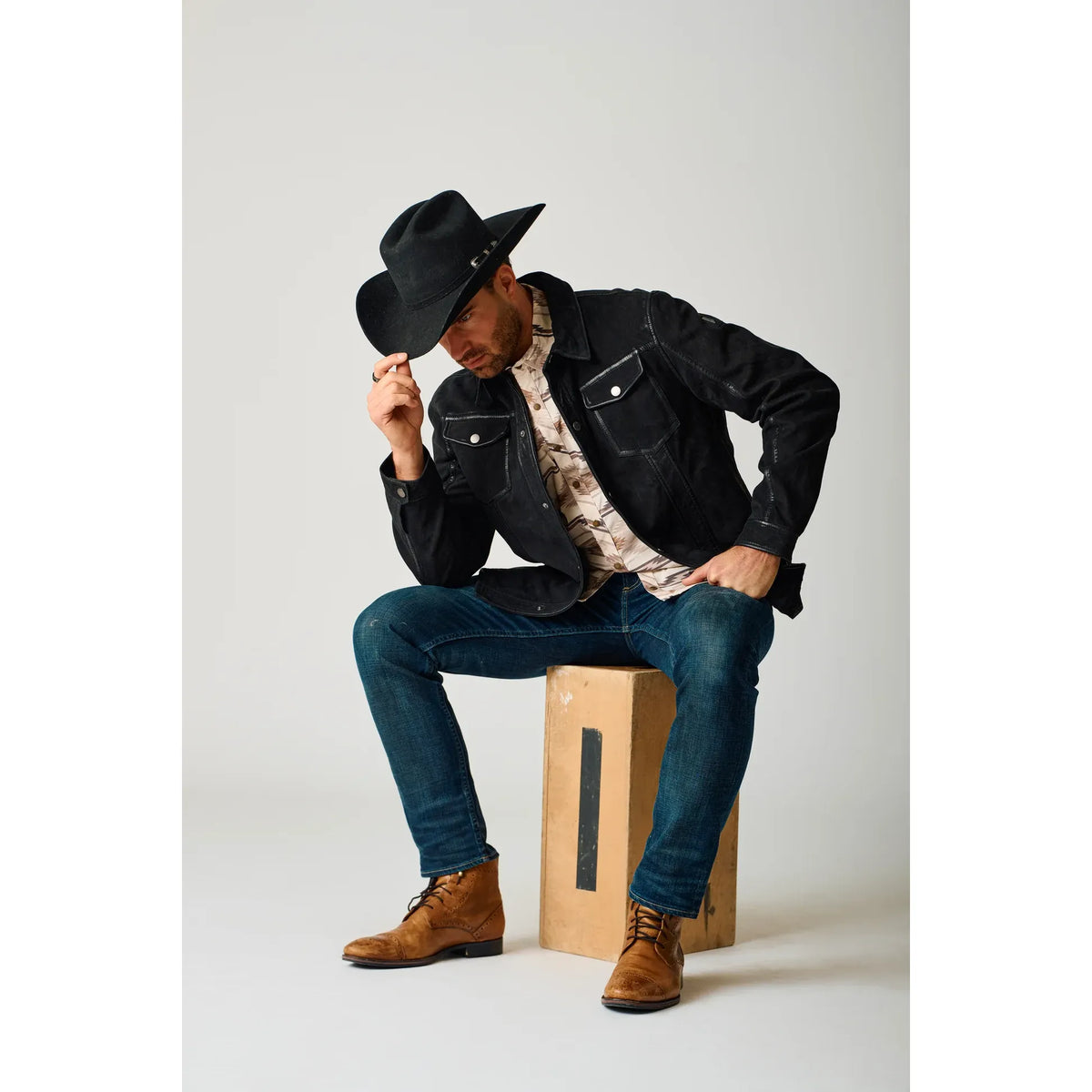 Man wearing Mauritius Sedro UF Nubuck Leather Jacket, camo shirt, jeans, and brown boots, tipping a black hat.