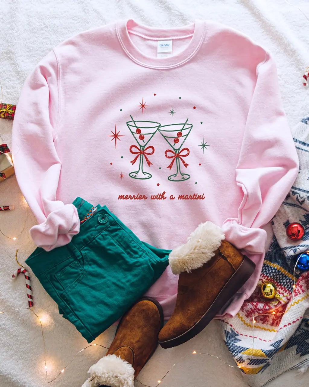 Light pink holiday crewneck featuring two clinking martini glasses with red bows and "merrier with a martini" text.