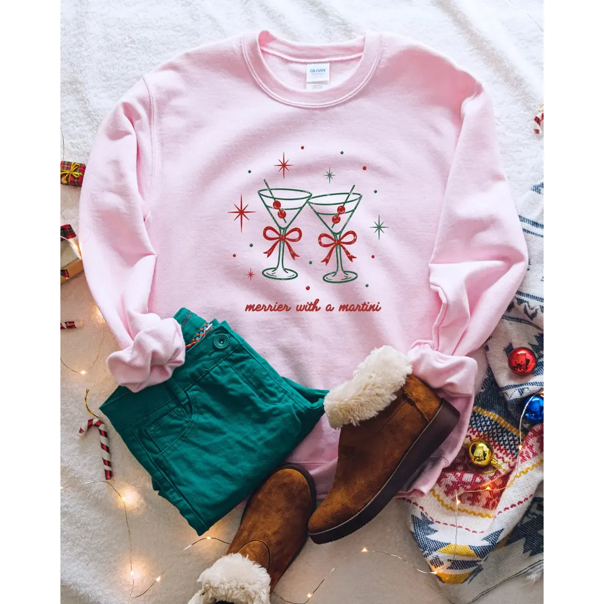 Light pink holiday crewneck featuring two clinking martini glasses with red bows and "merrier with a martini" text.