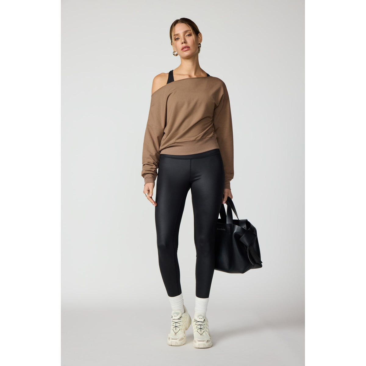 Model wears a brown flashdance sweatshirt with raw edge neckline and off-shoulder detail, paired with black leggings and white sneakers.