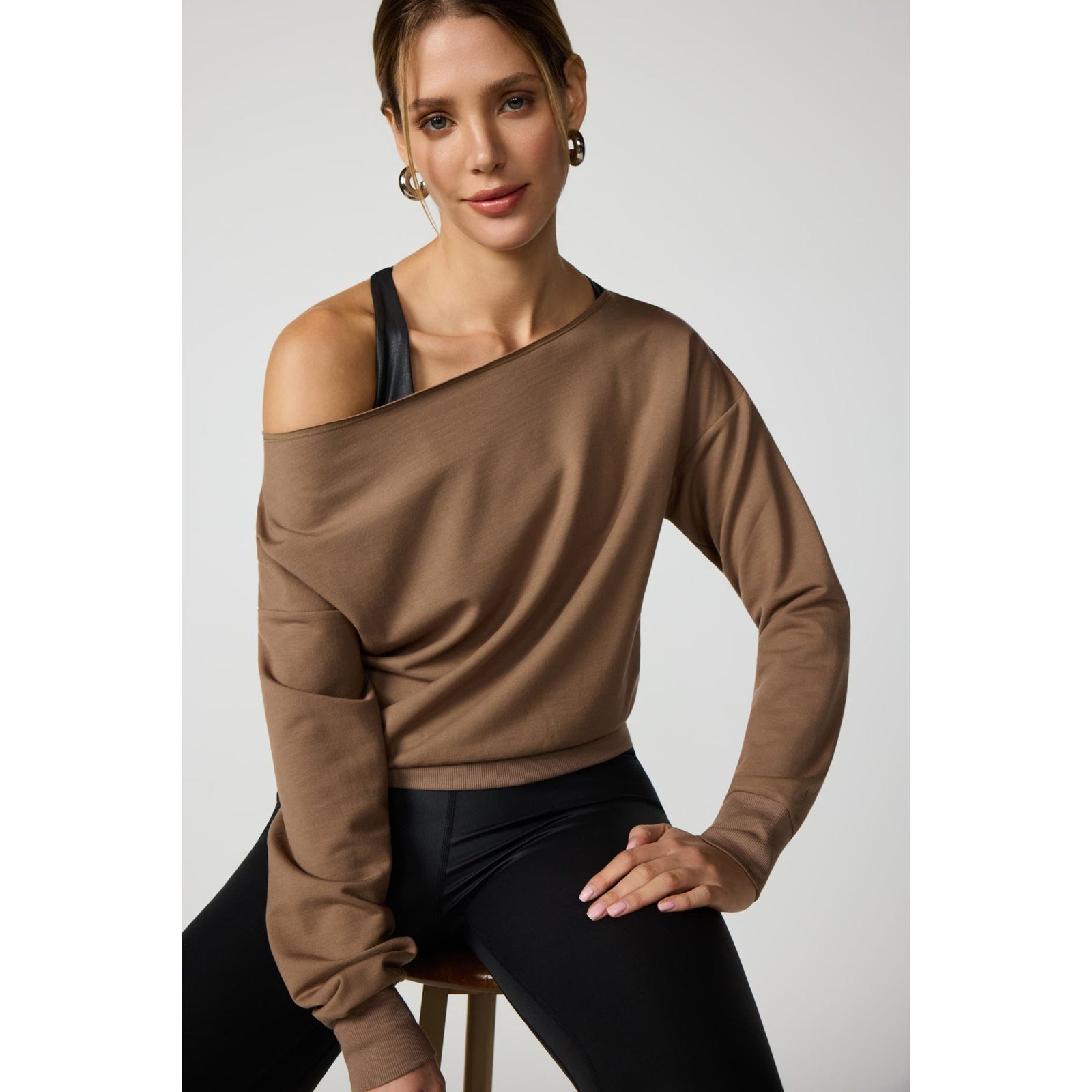 Brown off-shoulder sweatshirt with raw edge neckline, ribbed cuffs, and hem worn over black athletic tank top.